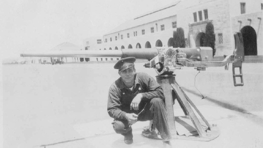 Marine in work uniform kneels next to a Sea School Gun